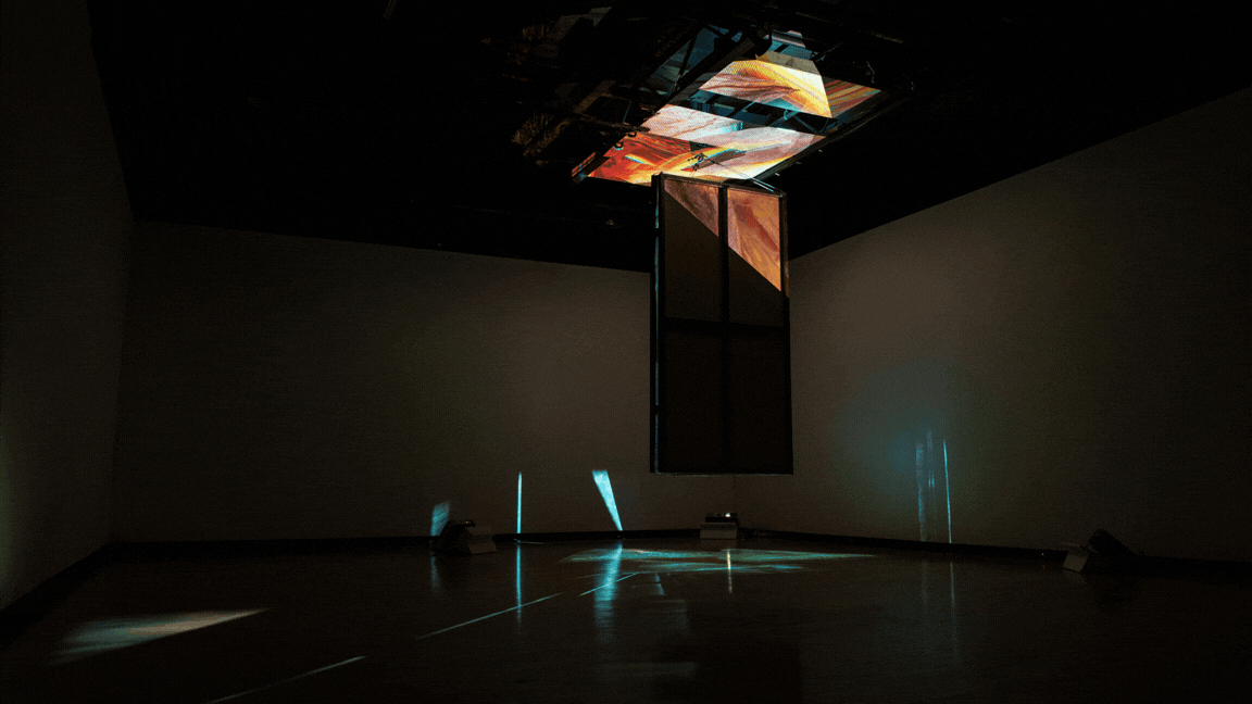 A suspended kaleidoscope structure hangs from the ceiling in a dark gallery, projecting warm orange and cool blue light shafts across the floor and walls.