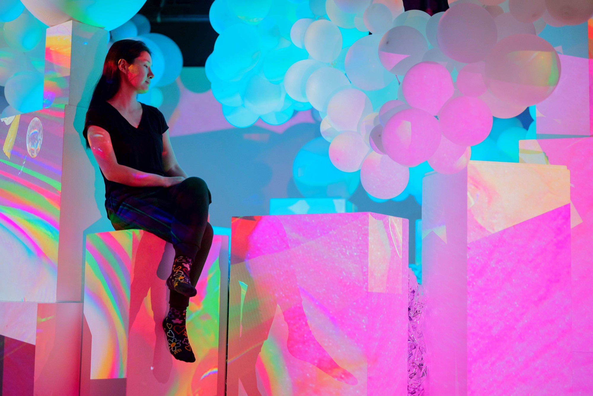 Bin Youn seated among illuminated white and pink balloon clusters and holographic panels, lit in cyan and magenta.