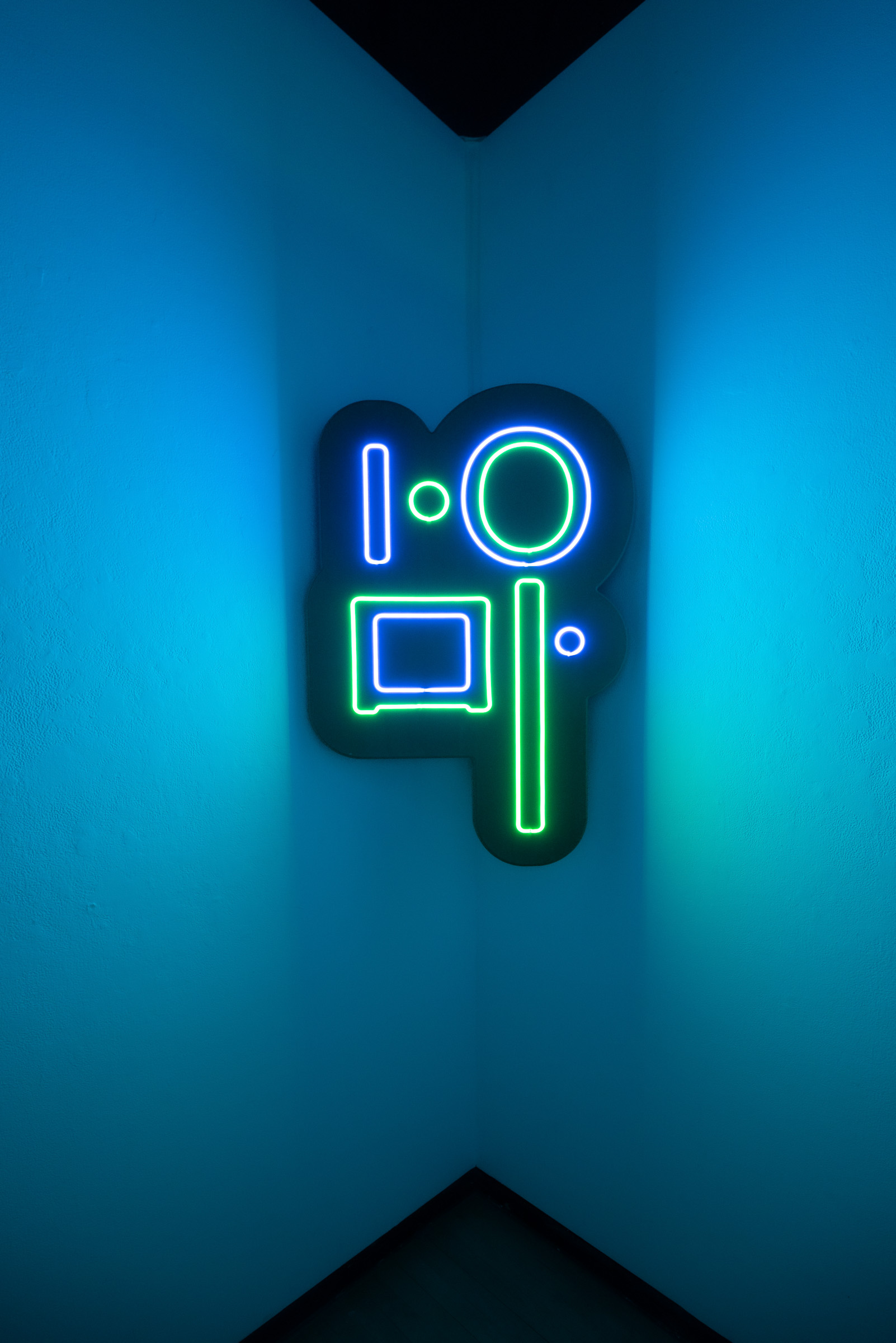 An abstract neon light sculpture in blue and green, its rounded and rectangular forms suggesting a stylized camera, glowing in a corner lit blue.