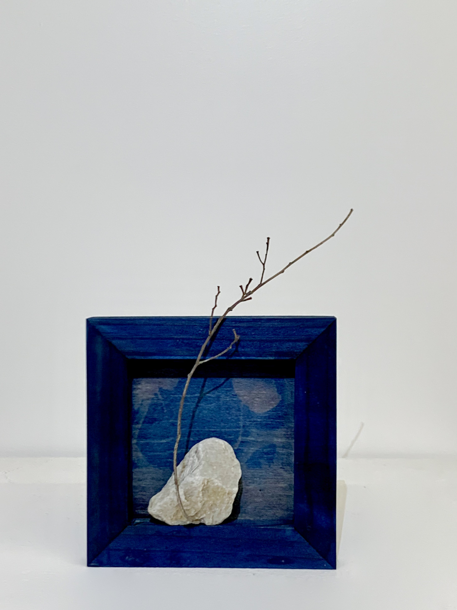 A small dark wooden frame containing a white stone and a bare branch, set against a cyanotype-blue ground, displayed on a gallery wall.