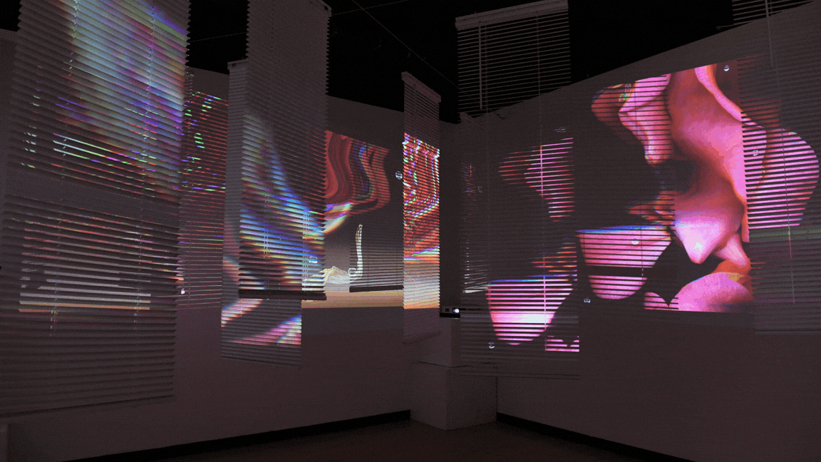 Suspended vinyl blinds cross the ceiling of a dark room, each strip lit with colorful projected imagery in pink and green tones.