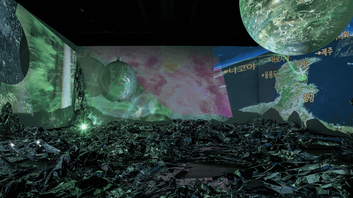 A large sphere resembling a planet, formed from projected video, floats above a gallery floor scattered with dark crumpled foil, lit in pink and green.