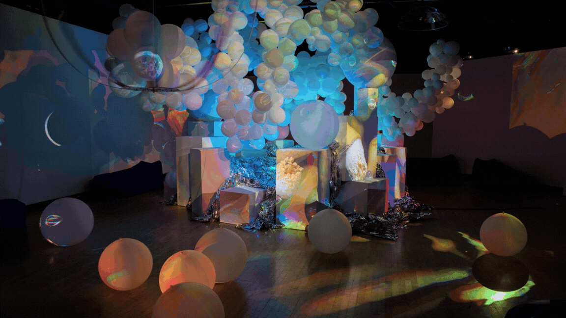 Large translucent spheres and geometric blocks catch swirling projected color patterns in a dim room, reflecting on the floor.