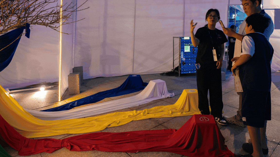 Installation view of Sacred Tree: four color-coded fabric trails, blue, white, yellow, and red, radiate across the floor from small covered platforms toward a bare branch hung with blue cloth, while the artist explains the piece to two young visitors holding phones.
