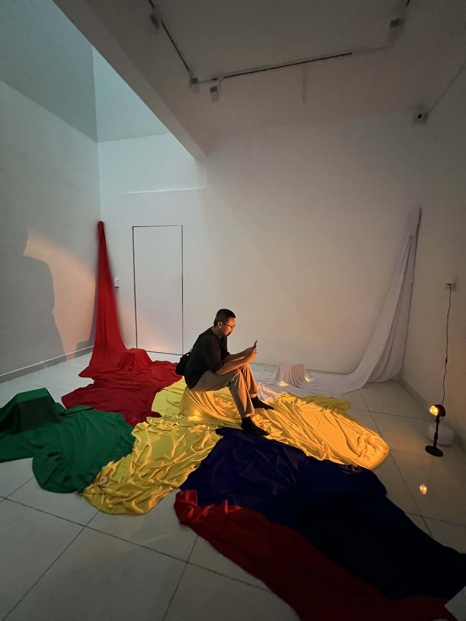 A visitor sits on a stool draped in yellow fabric at the centre of the installation, reading their phone, surrounded by six-metre fabric trails in red, green, yellow, and blue converging across a white gallery floor, a small warm lamp glowing at the edge.
