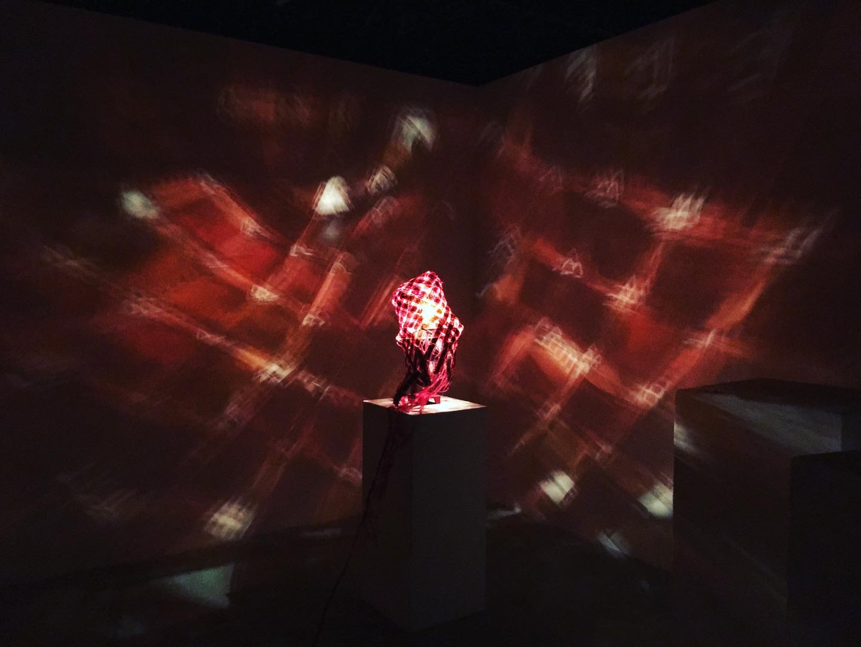 A woven red lamp shade glowing on a pedestal, casting a red patterned light across the surrounding walls in a dark room.